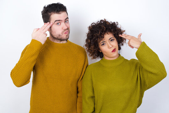 Unhappy Young Couple Wearing Knitted Sweater Standing Against White Wall Imitates Gun Shoot Makes Suicide Gesture Keeps Two Fingers On Temples.