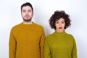 Joyful Young couple wearing knitted sweater standing against white wall looking to the camera, thinking about something. Both arms down, neutral facial expression.