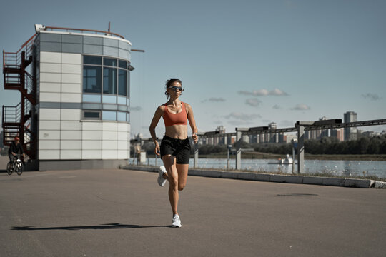 Full body of energetic motivated fit female in activewear with fitness tracker jogging on paved embankment near river in city