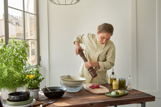Focused female chef in uniform adding seasonings with pepper mill to raw beef and mustard while cooking appetizing Steak tartare at home