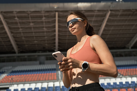 Low angle of modern fit female athlete in sportswear and trendy sunglasses with smartwatch browsing mobile phone while standing on stadium tribune and resting after training