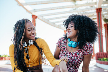 Cheerful black female friends with braids and curly hair sitting on embankment and having fun in summer