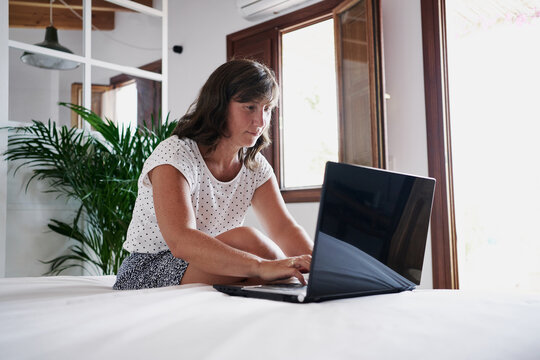 Side View Of Serious Adult Female Freelancer Sitting On Bed In Room And Working On Remote Project While Using Netbook