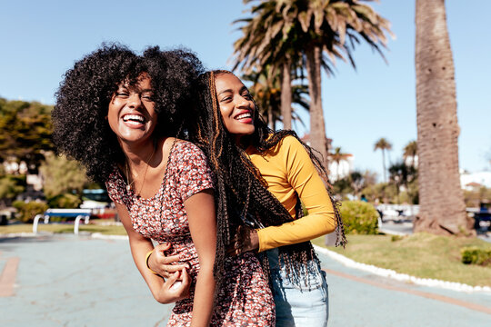Side view of black woman with curly hair hugging delighted African American female friend with braids while standing on embankment and looking away