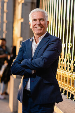 Happy gray haired senior executive male in elegant blue suit standing with arms crossed near metal fence and looking at camera