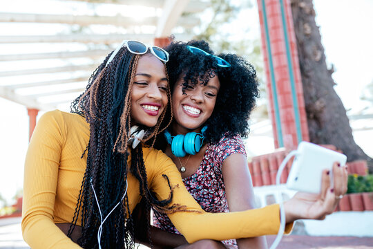 Smiling black woman with braids hugging cheerful African American female friend with curly hair and taking selfie together while standing on promenade in summer