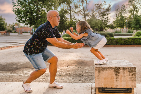 Side view of father standing in park and ready for catching playful daughter jumping from stone parapet while entertaining together in city