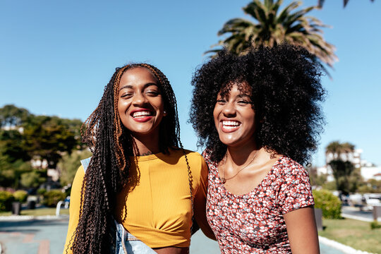 Delighted black woman with braids holding hand of African American female best friend with curly hair while walking along street in tropical city on sunny day looking at camera