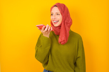 Smiling Young Caucasian Muslim woman wearing hijab standing against yellow wall sending voice message on her smart phone. Communication and new technologies concept.