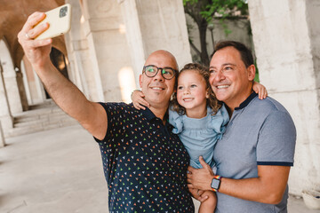Delighted gay couple with cute daughter standing on street and taking selfie on smartphone during stroll in summer