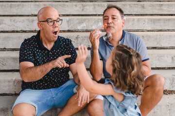 Side view of cheerful gay couple with cute little girl having fun with soap bubbles while spending time together in park
