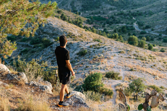 Back view of anonymous young male with hiking poles standing on mountain slope and enjoying landscape while taking break during trekking workout