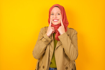 Strong healthy straight white teeth. Close up portrait of happy Young caucasian Muslim woman wearing hijab standing against yellow wall with beaming smile pointing on perfect clear white teeth.