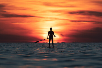 Silhouette of faceless female surfer standing on paddle board and rowing against spectacular sun in sunset sky