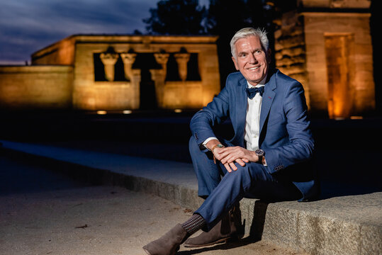 Positive elegant senior male in stylish blue suit with bow tie and wristwatches sitting on stone border in night city and looking away