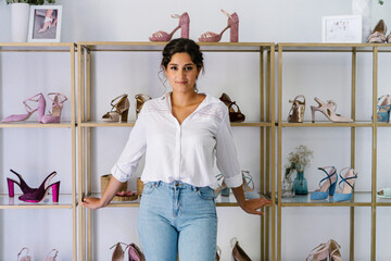Charming female in casual outfit standing near wooden shelves with various high heeled shoes in expensive boutique and looking at camera