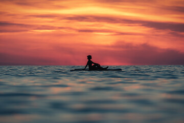 Silhouette of faceless female surfer sitting on paddle board and rowing against spectacular sun in sunset sky