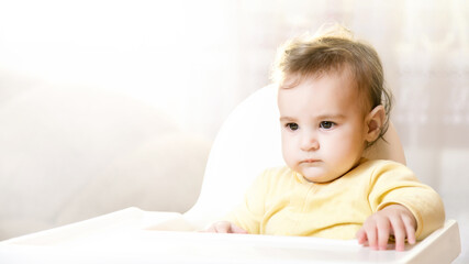 Funny portrait of a disgruntled toddler on a high chair indoors. Beautiful sad baby