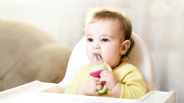 Little baby sitting in high chair and eating food indoors.. baby's first feeding. 6 month old toddler with nibbler in the mouth
