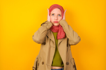 Frustrated Young caucasian Muslim woman wearing hijab standing against yellow wall plugging ears with hands does not wanting to listen hard rock, noise or loud music.