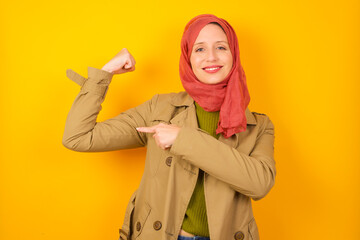 Smiling Young caucasian Muslim woman wearing hijab standing against yellow wall raises hand to show muscles, feels confident in victory, strong and independent.