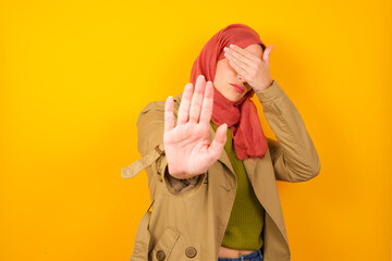 Young caucasian Muslim woman wearing hijab standing against yellow wall covers eyes with palm and doing stop gesture, tries to hide. Don't look at me, I don't want to see, feels ashamed or scared.