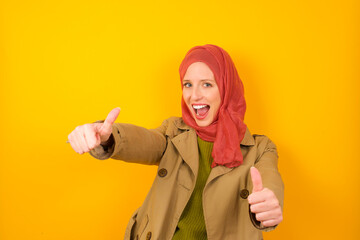 Young caucasian Muslim woman wearing hijab standing against yellow wall making positive gesture with thumbs up smiling and happy for success. Looking at the camera, winner gesture.