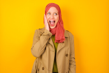 Young caucasian Muslim woman wearing hijab standing against yellow wall excited looking to the side hand on face. Advertisement and amazement concept.