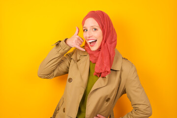 Young caucasian Muslim woman wearing hijab standing against yellow wall imitates telephone conversation, makes phone call gesture with hands, has confident expression. Call me!