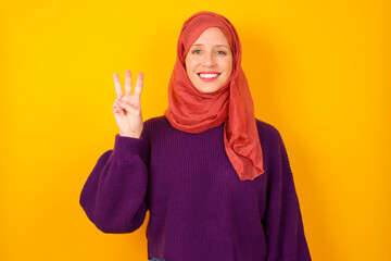 Young caucasian Muslim woman wearing hijab standing against yellow wall showing and pointing up with fingers number three while smiling confident and happy.