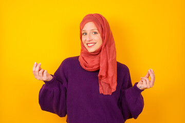 Young caucasian Muslim woman wearing hijab standing against yellow wall, making money gesture.