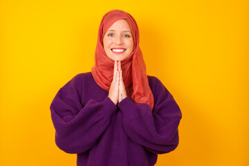 Young caucasian Muslim woman wearing hijab standing against yellow wall keeps palms together, has...