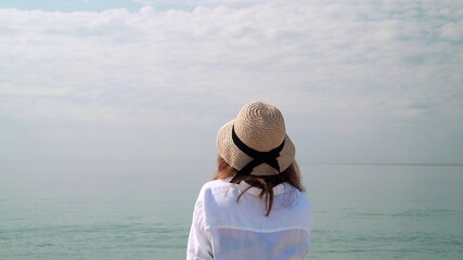 Young woman in hat enjoying horizon, water and sky. Back of woman in white blouse and wicker hat on background of water, ocean and cloudy sky