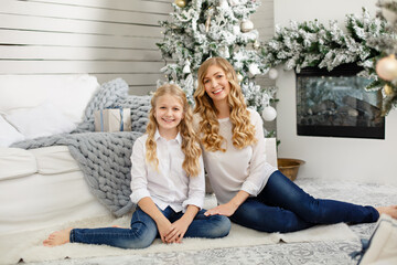 Obraz premium Girl pre-teen with her mpther near fir-tree smile to the camera. Christmas celebration. Daughter with Mom weared white shirts and denim. New year eve. Happy family time. Light interior with fireplace