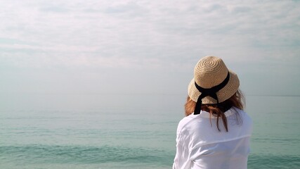 Young woman in hat enjoying horizon, water and sky. Back of woman in white blouse and wicker hat on background of water, ocean and cloudy sky