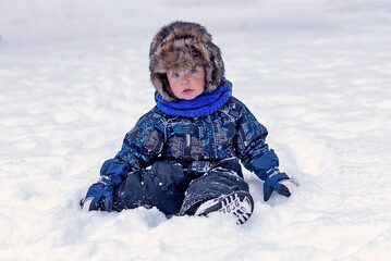 Fototapeta na wymiar Funny little boy in overalls and fur hat clothes playing outdoors during snowfall. Active rest with children in winter on cold snowy days. Happy child.