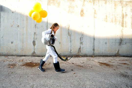 Little Girl Walking With Space Helmet And Holding Balloon On Street During Sunny Day