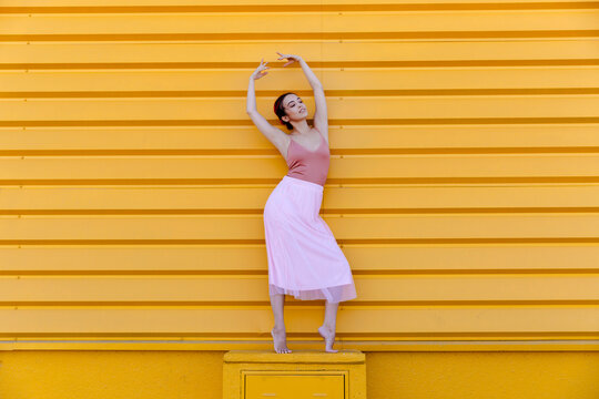 Ballerina with arms raised posing while standing on seat against yellow wall