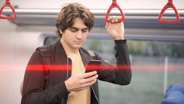 Man holding by red handrails in train, man typing in phone on background of white train car. Middle shot of man in black jacket holding a phone, inside public transport