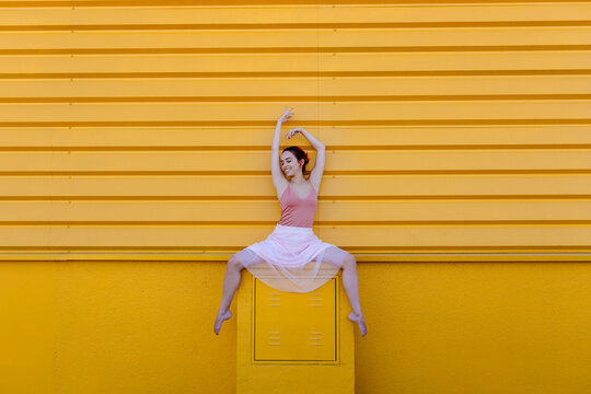 Smiling ballerina with arms raised dancing on seat against yellow wall