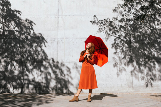 Smiling woman holding umbrella while standing against tree shadow wall