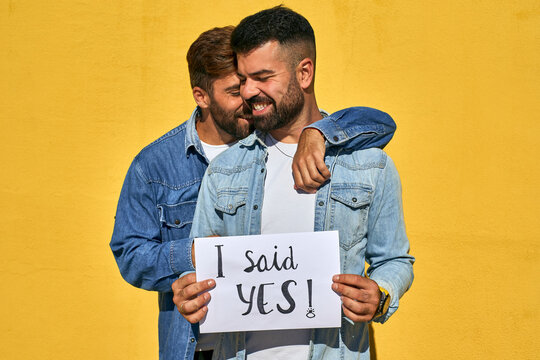 Gay man embracing young man holding paper with text I Said Yes written on it against yellow wall