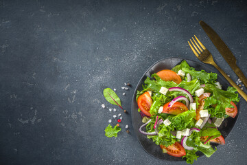 Fototapeta na wymiar Bowl of healthy vegetable salad with various leaf salad, tomatoes, feta cheese, onions, olive oil, dark grey concrete background copy space