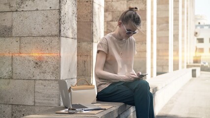 Young woman learning outside with phone in hand near university building, lens flare. Young student in sunglasses typing in the phone, outdoors