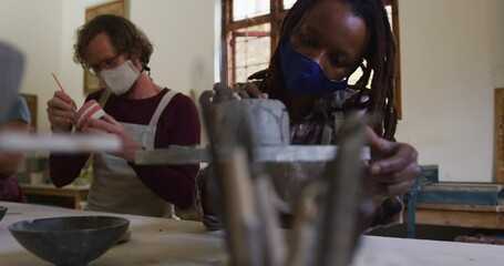 Diverse male and female potters wearing face mask and apron working on pottery at pottery studio - Powered by Adobe