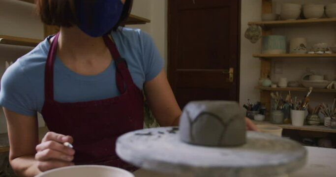 Female caucasian potter wearing face mask and apron using ribbon tool to create designs on pot at po