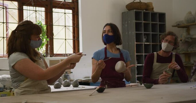 Diverse potters wearing face masks and aprons painting pots at pottery studio