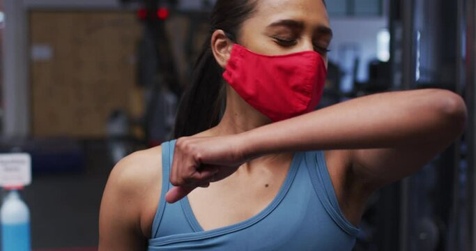 Fit caucasian woman wearing face mask sneezing on her elbow in the gym