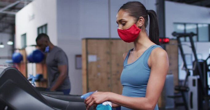 Fit caucasian woman wearing face mask cleaning treadmill machine with disinfectant in the gym