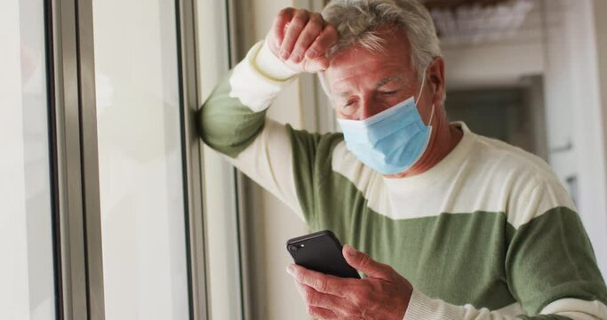 Senior caucasian man wearing face mask using smartphone near window at home